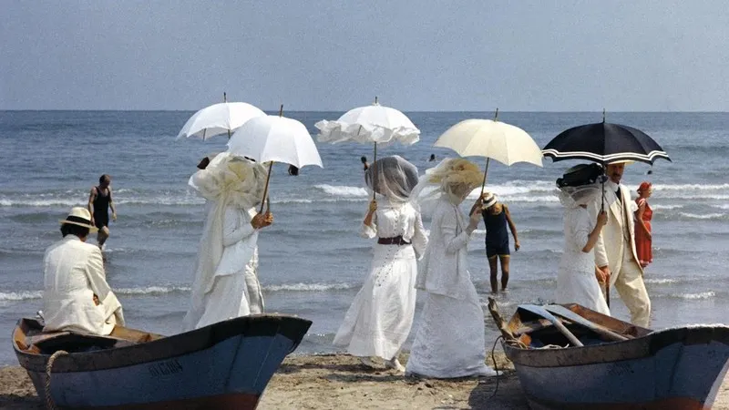 Fotograma de Morte a Venezia, de Luchino Visconti. Mostra unha escena na praia, ambientada a finais do século XIX, cun grupo de mulleres vestidas de branco e con parasoles xunto ao progatonista, de traxe branco, nunha esquina, sentado sobre unha barquiña. Todos están de costas á cámara e, de fondo, o mar.
